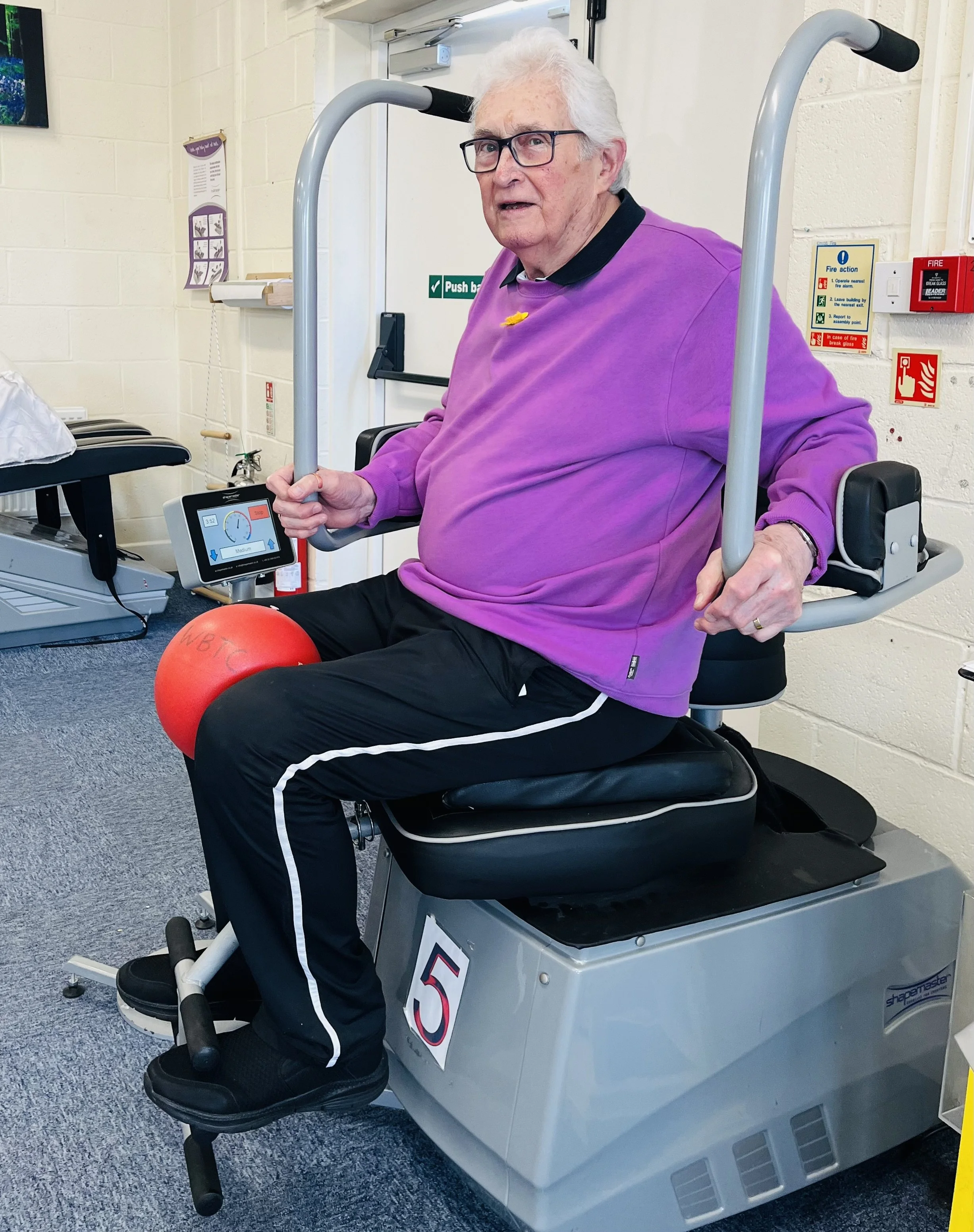 An elderly man with white hair and glasses exercising on a mechanical lift chair with handles in a therapy room. He is wearing a purple sweatshirt, black pants with white stripes, and black shoes, with a red exercise ball on his lap.