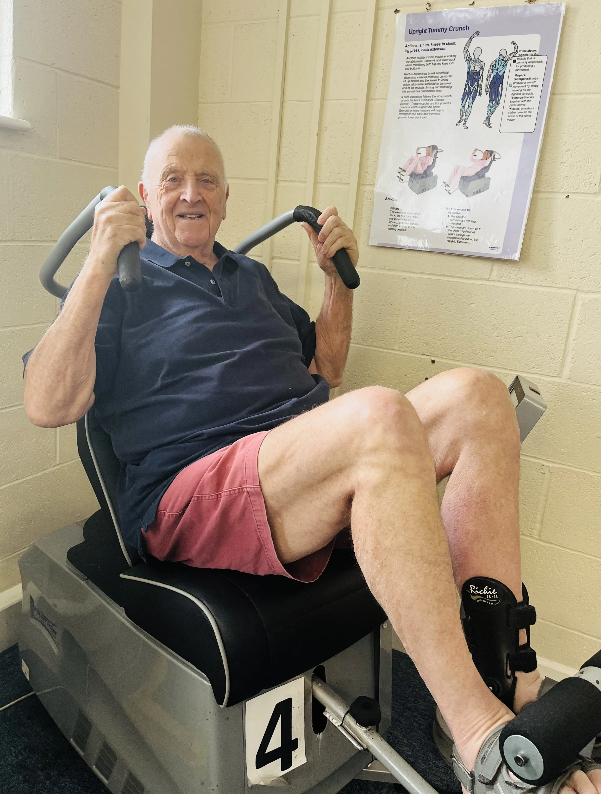 An elderly man with a happy expression, sitting on a medical exercise machine, holding onto the handles, wearing a navy shirt and pink shorts. There is a poster with exercise instructions on the wall behind him.