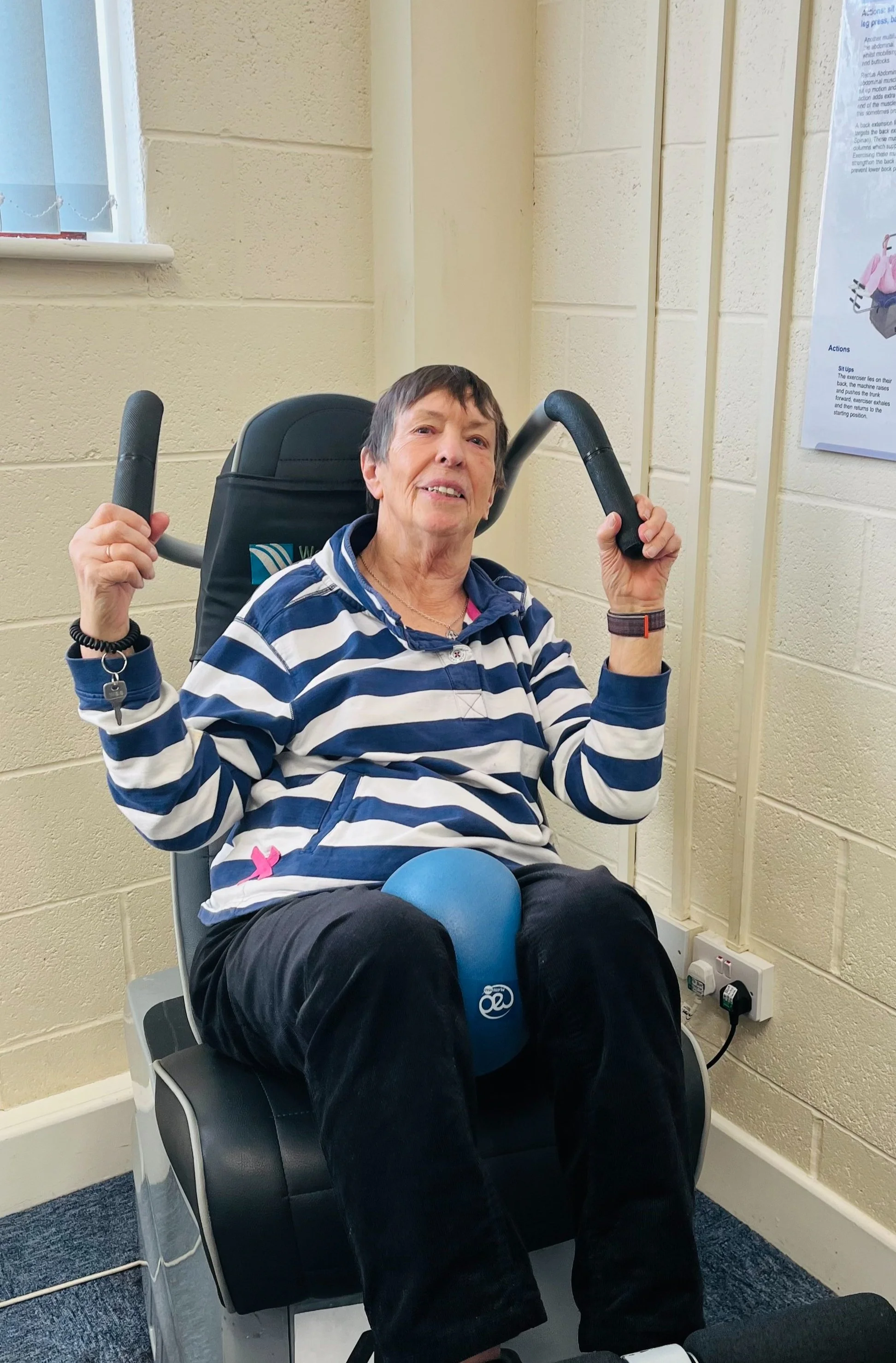 An elderly woman in a striped blue and white shirt exercises on a seated device with handles and a blue exercise ball resting on her lap. She is indoors against a beige brick wall, with a window to her left and a poster on the wall to her right.