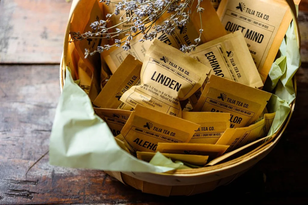 A woven basket containing various paper tea packets from Tilia Tea Co., including Linden, Echinacea, and other herbal teas, with dried lavender flowers on top and lined with light green paper, placed on a wooden surface.