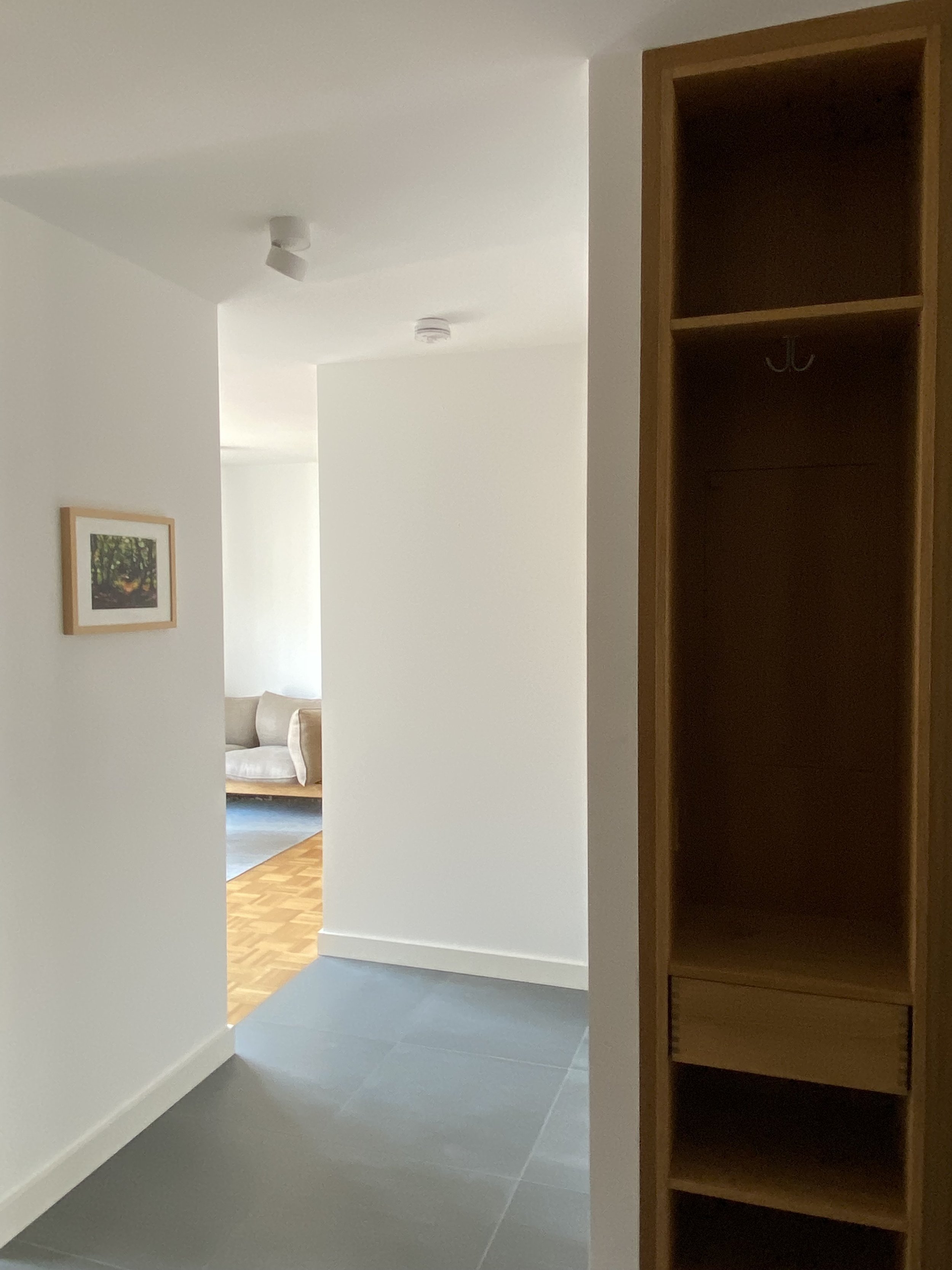 Interior of a modern apartment with white walls, a wooden shelf, a wall painting, and a view into a room with a light-colored sofa and parquet flooring.