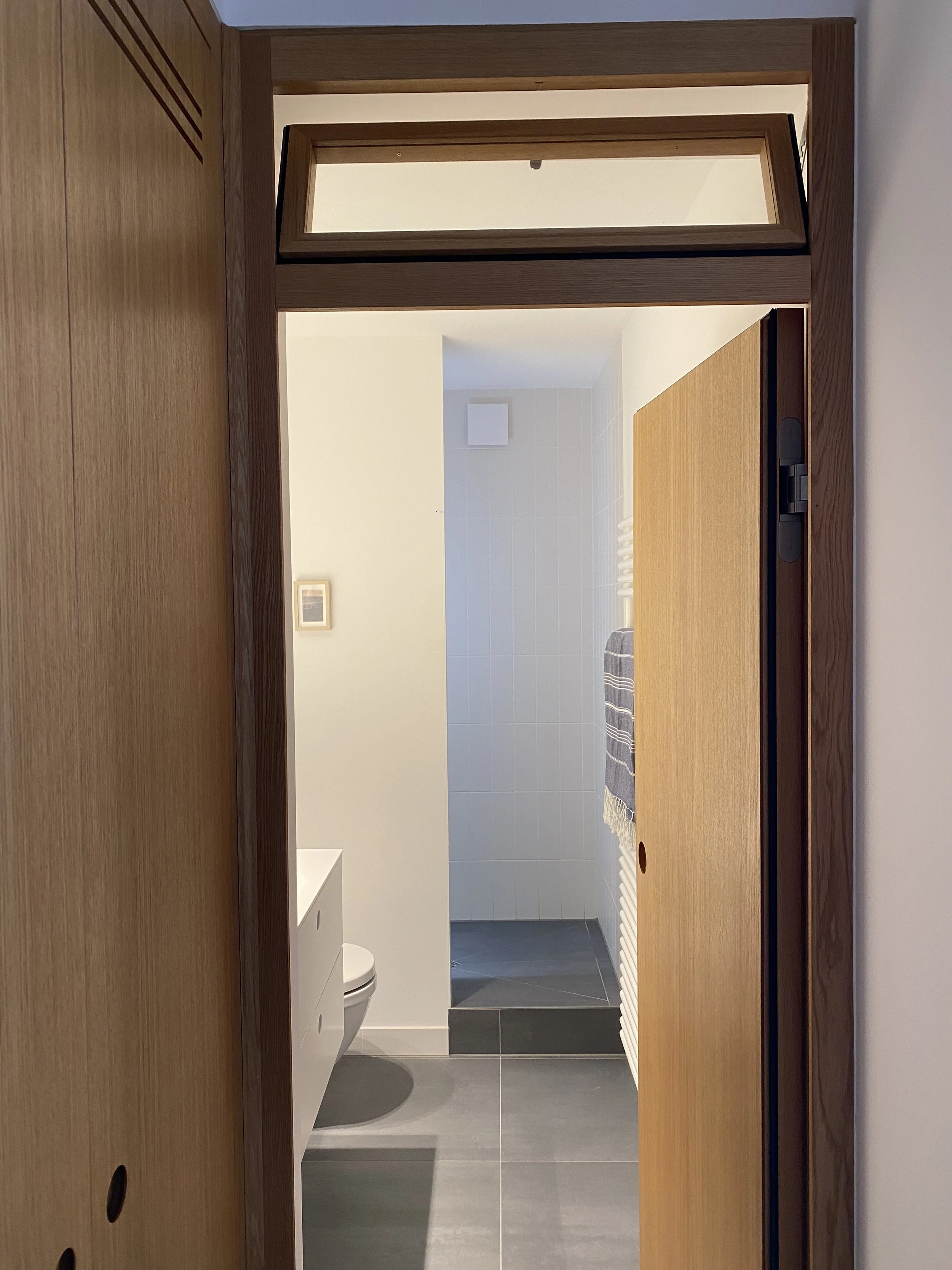 Interior of a modern bathroom with an open wooden door, dark floor tiles, a white wall, a towel rack with a striped towel, and a wall-mounted toilet.