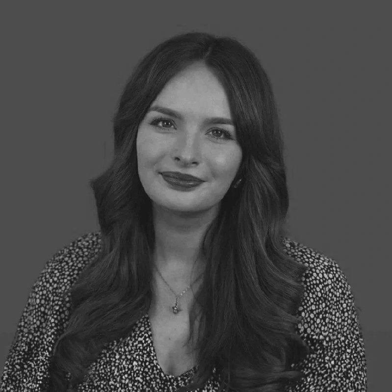 Black and white portrait of a young woman with long wavy hair, wearing a patterned blouse and a necklace, smiling gently against a plain background.