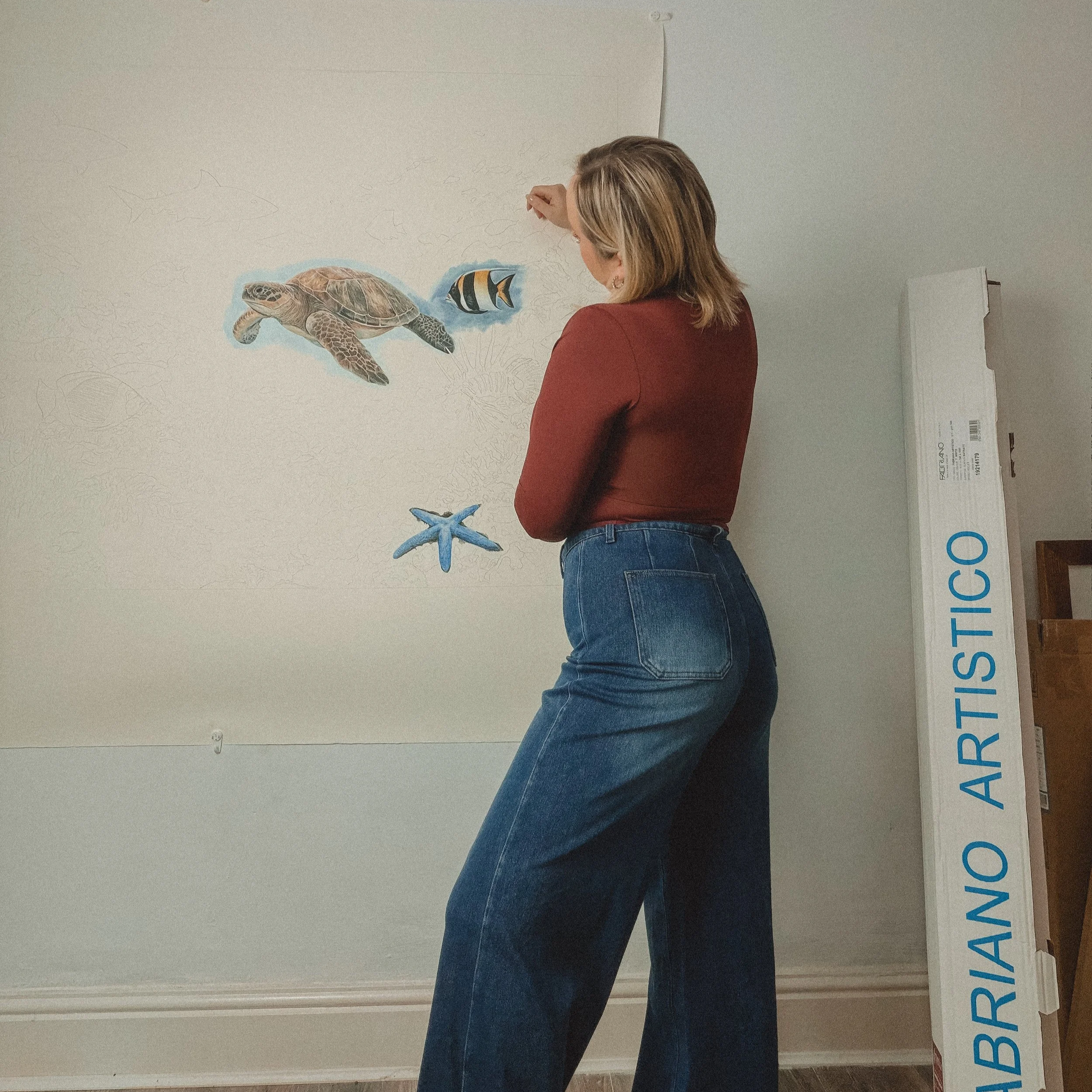 fine artist in front of a mural with marine animals, including a turtle, fish, starfish, and sharks, on a white wall. all hand drawn