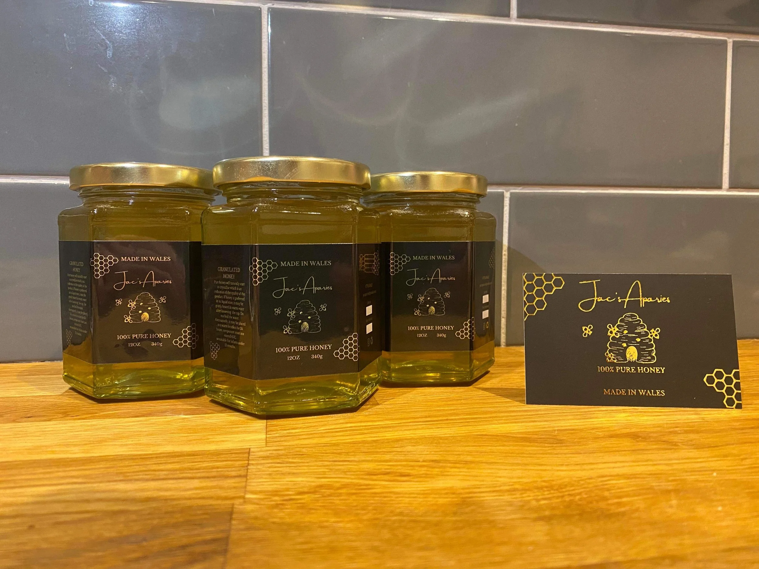 Three jars of honey and a business card on a wooden surface. The jars have black labels with gold accents and text, and are sealed with gold lids. The business card matches the jar labels in color and design, featuring honeycomb patterns, a honey jar