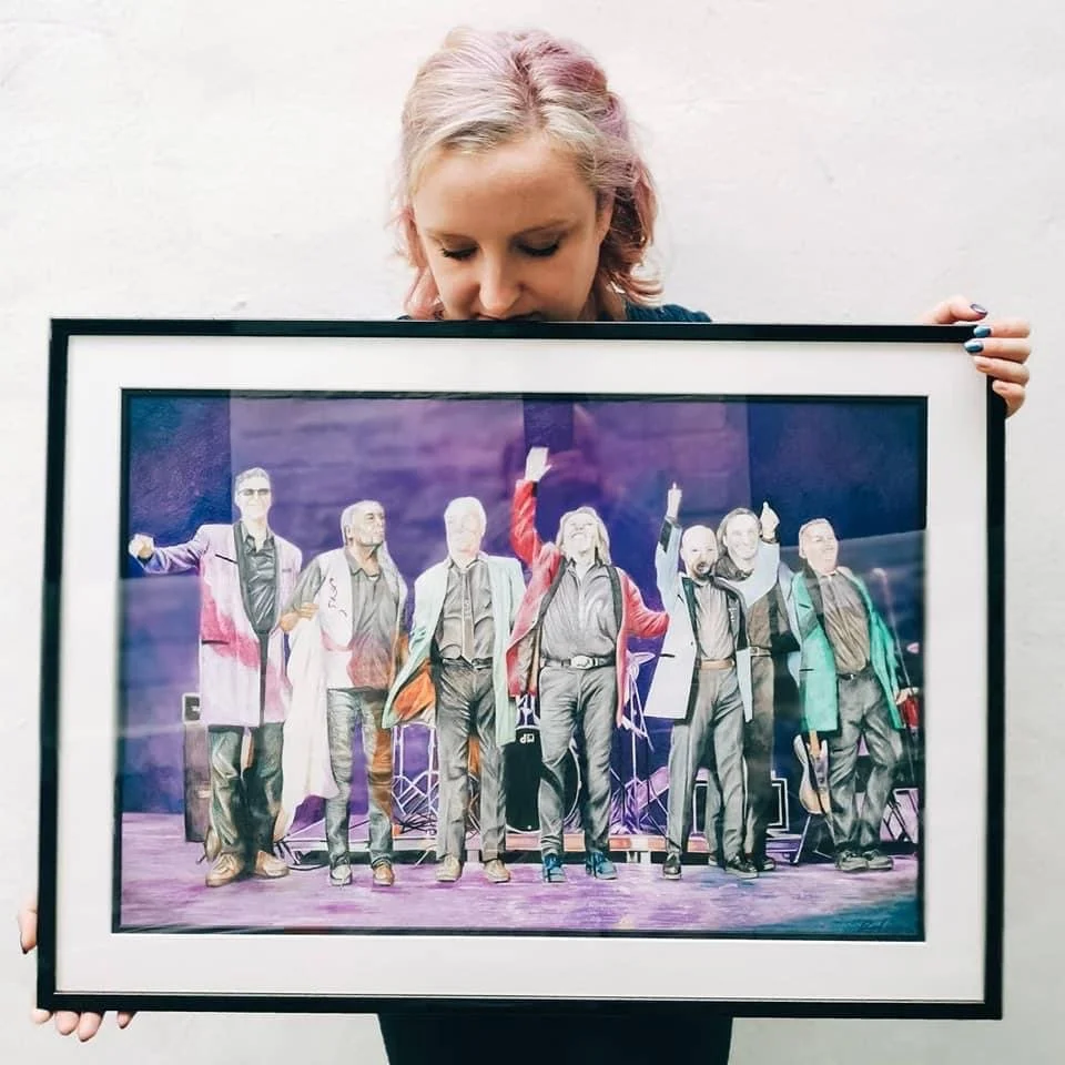 A woman with pink and blonde hair holding a framed photo of seven people on stage.