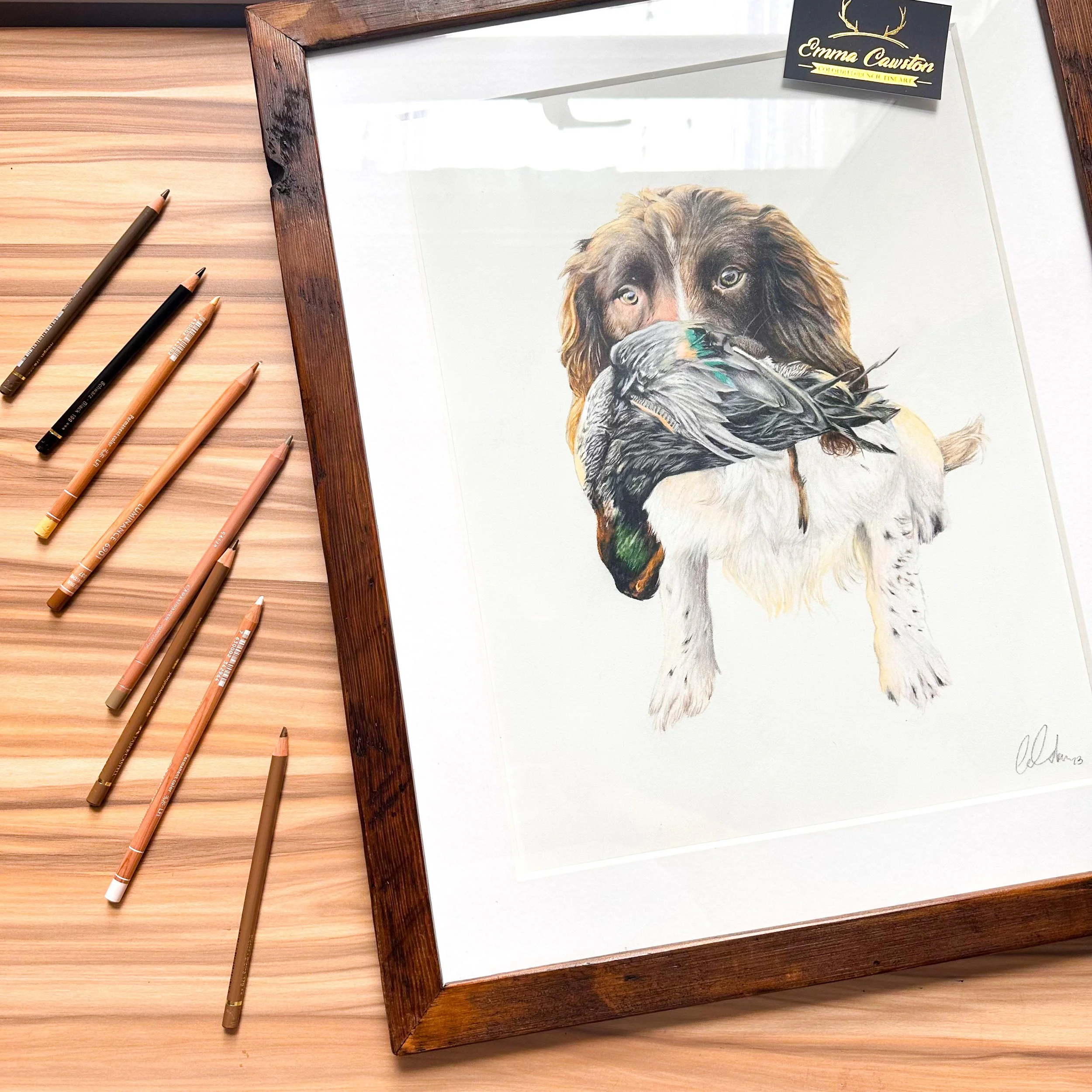 A framed colored pencil drawing of a dog with a bird in its mouth, on a wooden surface with colored pencils beside it.