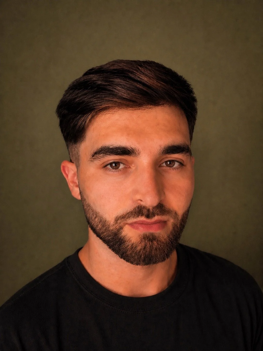 A portrait of a man with dark brown hair, a full beard, wearing a black shirt, standing against a plain olive-green background.