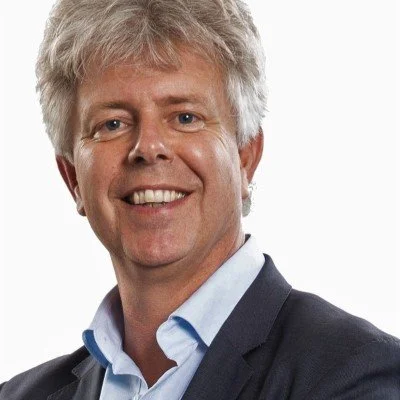 A middle-aged man with gray hair smiling, wearing a dark suit jacket and a light blue dress shirt, posing against a plain white background.