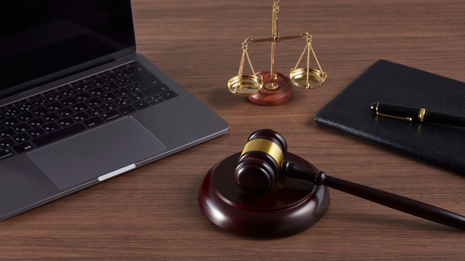 Legal office desk with a laptop, a wooden gavel on a sound block, a scale of justice, a black notebook with a gold-trimmed pen, all on a wooden surface.