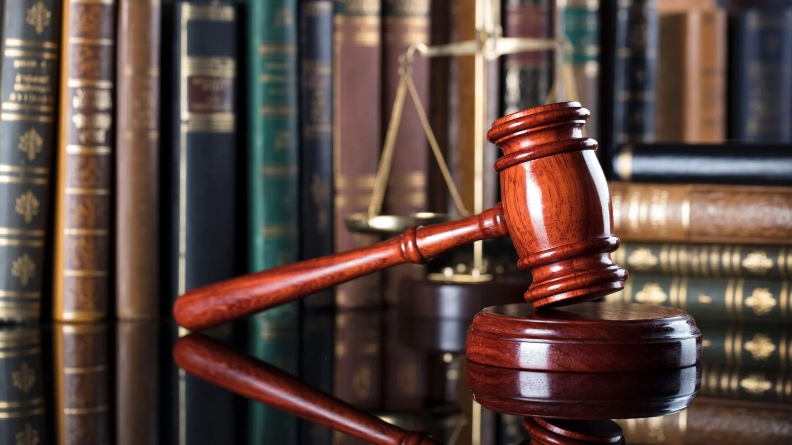 A wooden judge's gavel resting on a sound block with legal books in the background and a scale of justice in the setting.