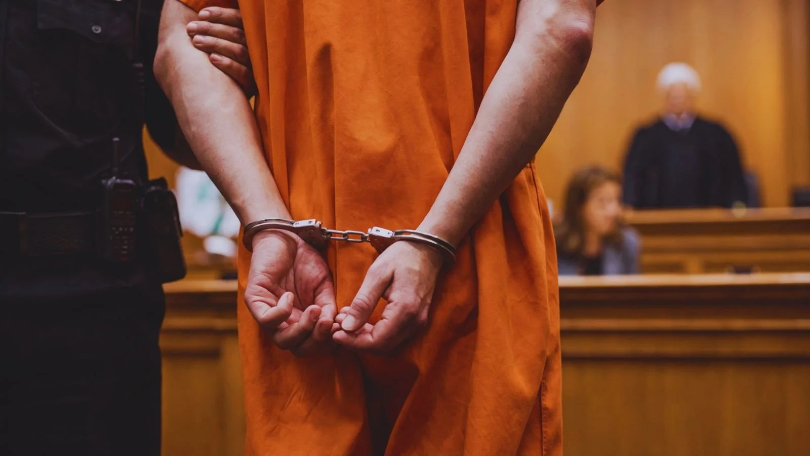 A person in an orange jumpsuit handcuffed in court, with a judge and a lawyer visible in the background.