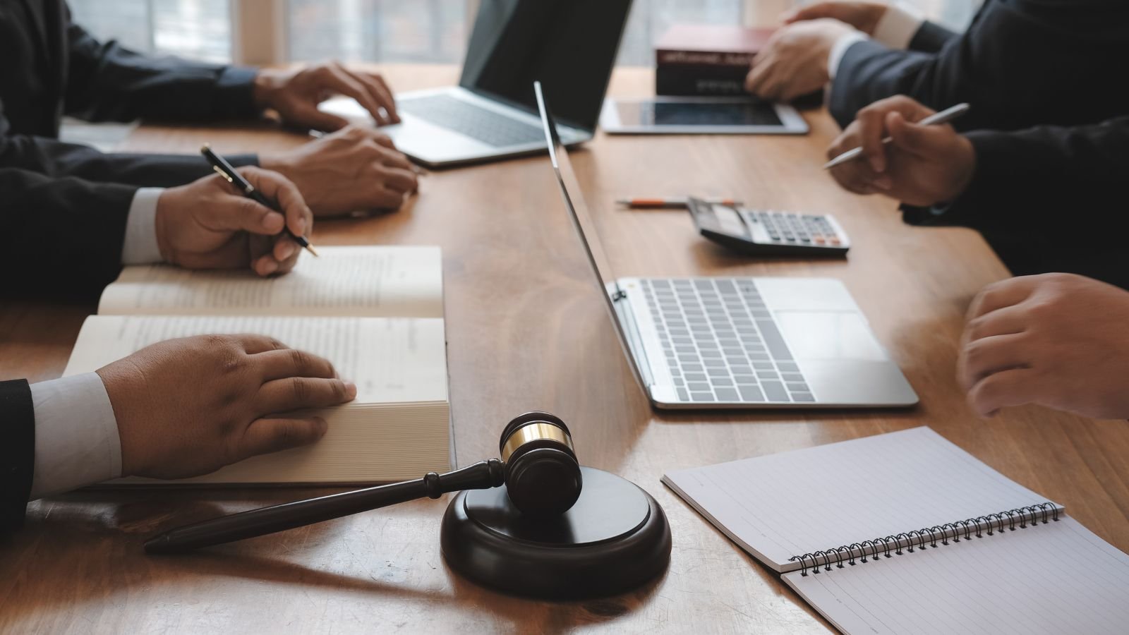 Business meeting with several people seated at a wooden table, with laptops, notebooks, a gavel, and writing instruments.