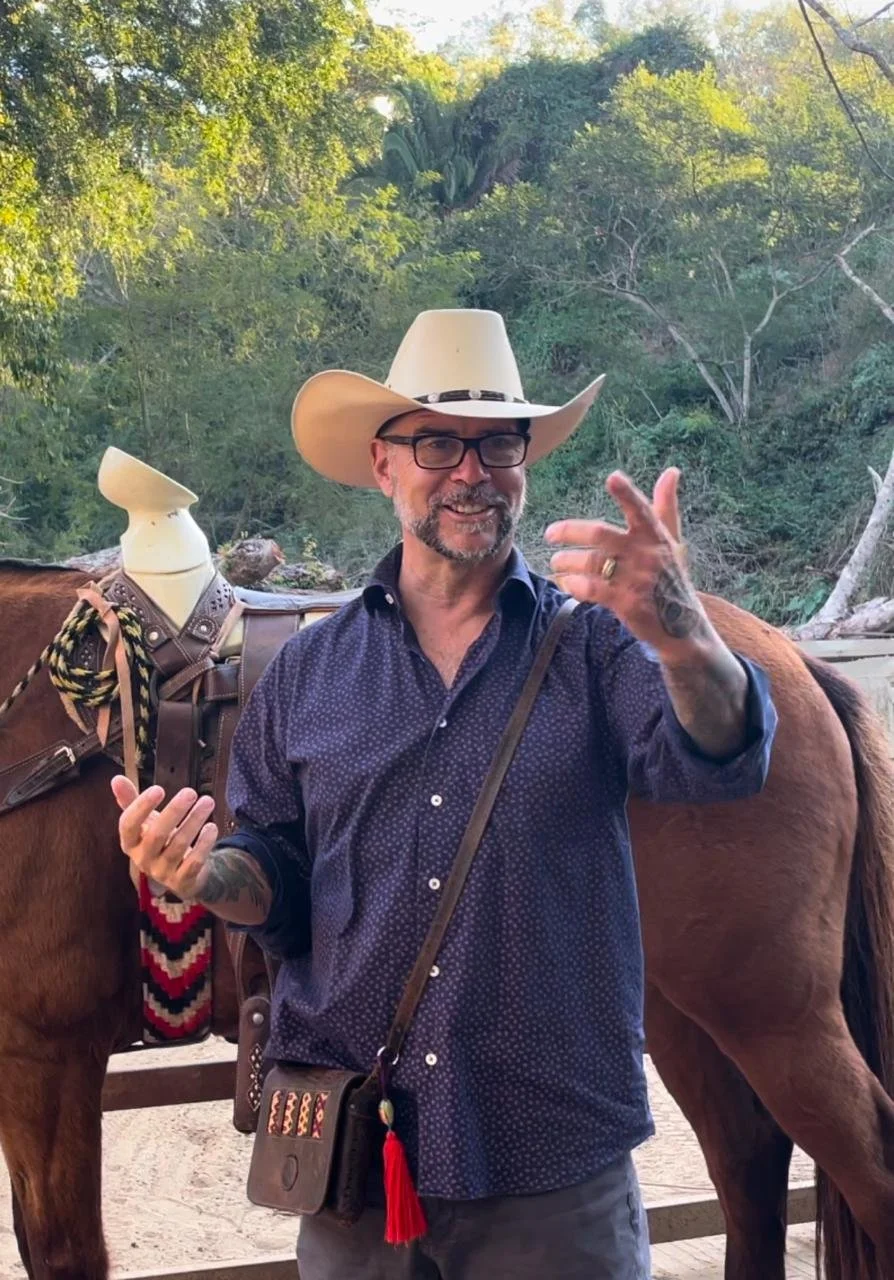 A man wearing glasses and a cowboy hat, smiling and gesturing with his hand, standing outdoors next to a horse with a saddle and colorful accessories, in a green forested area.