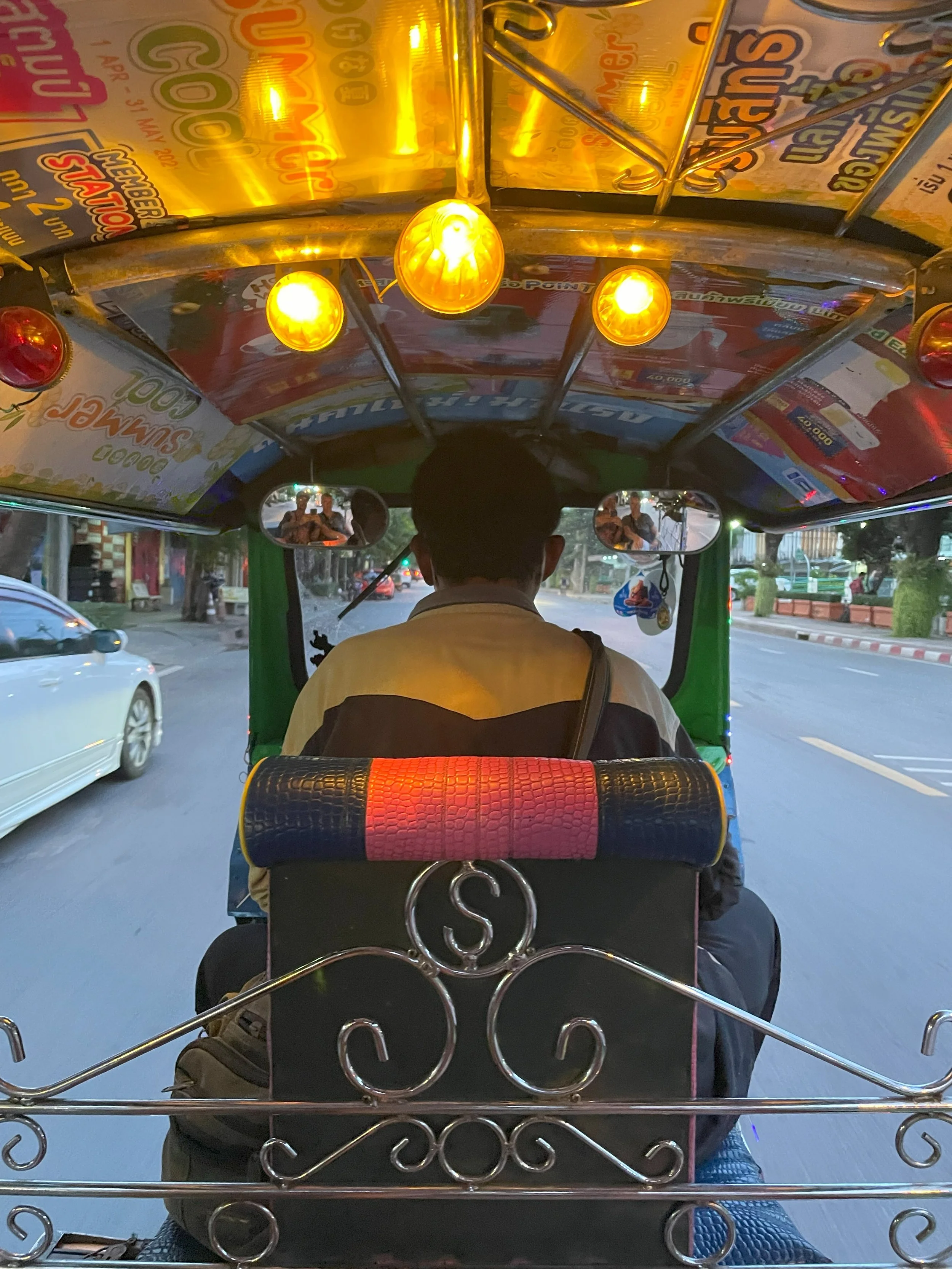 Inside a tuk-tuk taxi on a city street during dusk, with a driver in front and a person reflected in the side mirror, and colorful advertising posters on the vehicle's roof.