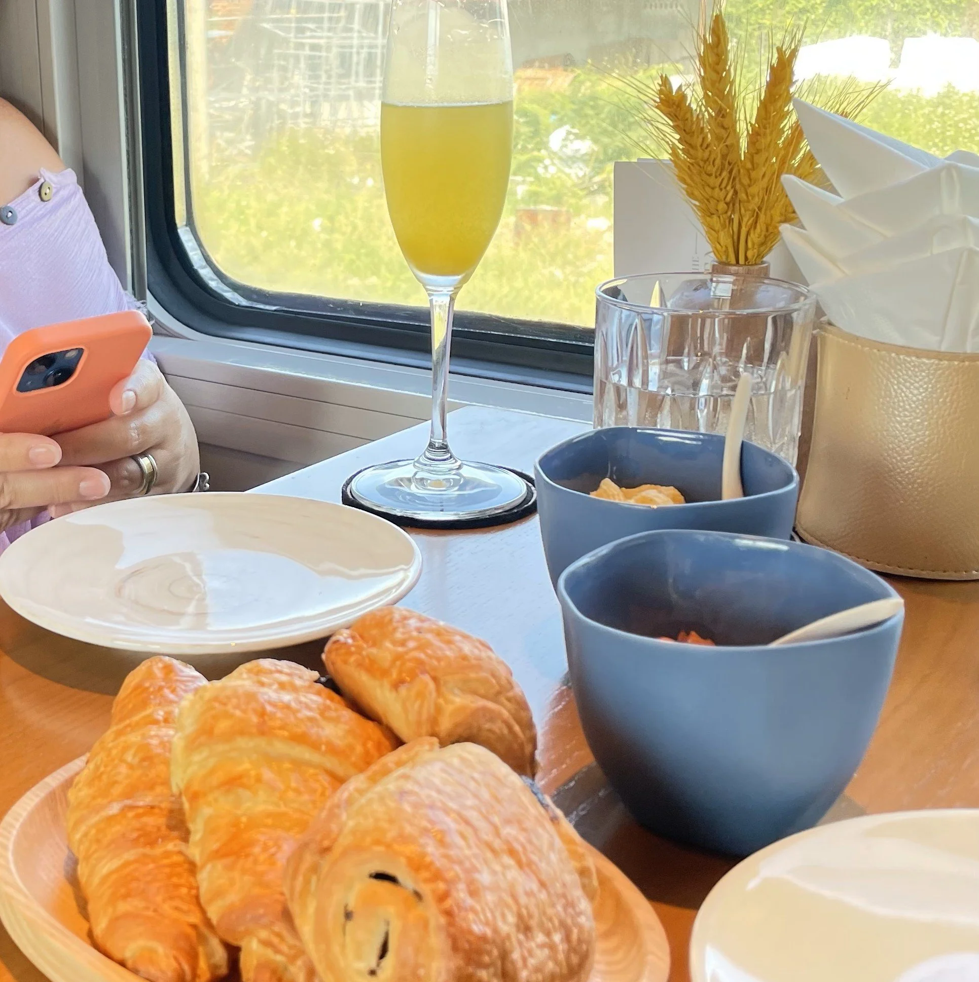 A breakfast table with croissants, a glass of yellow beverage, bowls of food, napkins, flowers, and a person using a smartphone near a window.