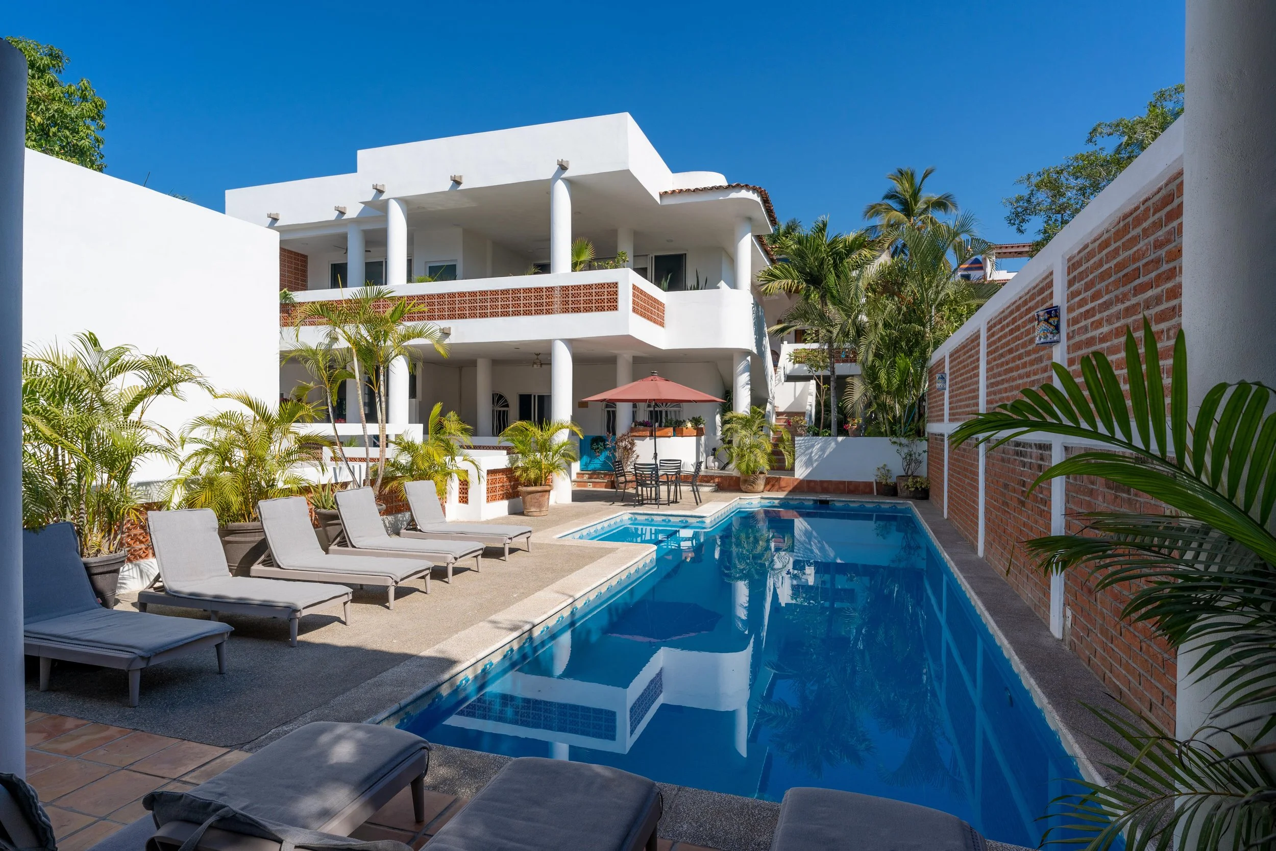 A swimming pool surrounded by lounge chairs and potted plants in a sunny outdoor courtyard of a white multi-story building with balconies, under a clear blue sky.