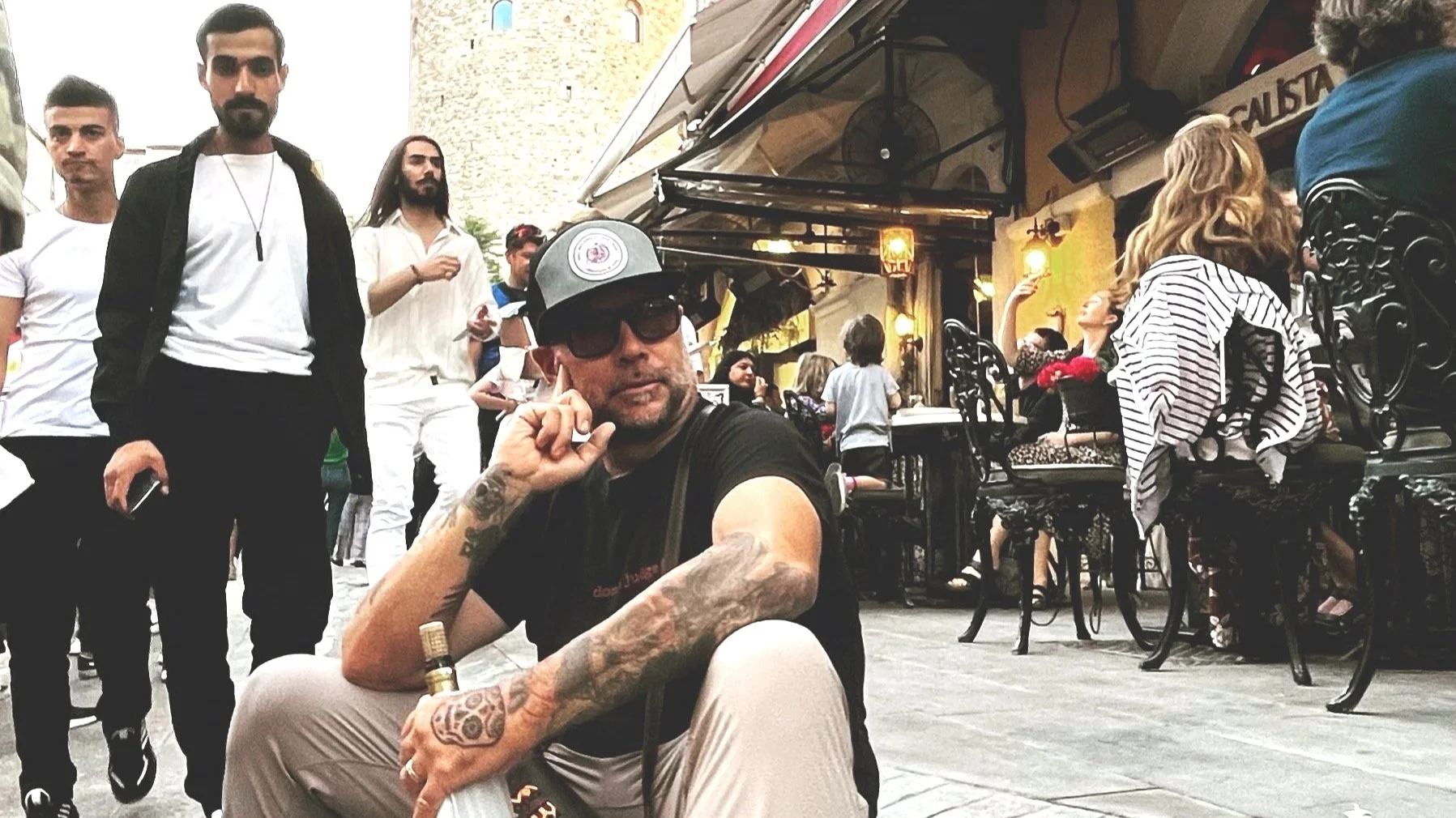 A man with tattoos, wearing sunglasses and a cap, sitting on the ground in a crowded outdoor cafe or street scene, with several people standing and sitting around, some looking at the camera and others engaged in conversation.