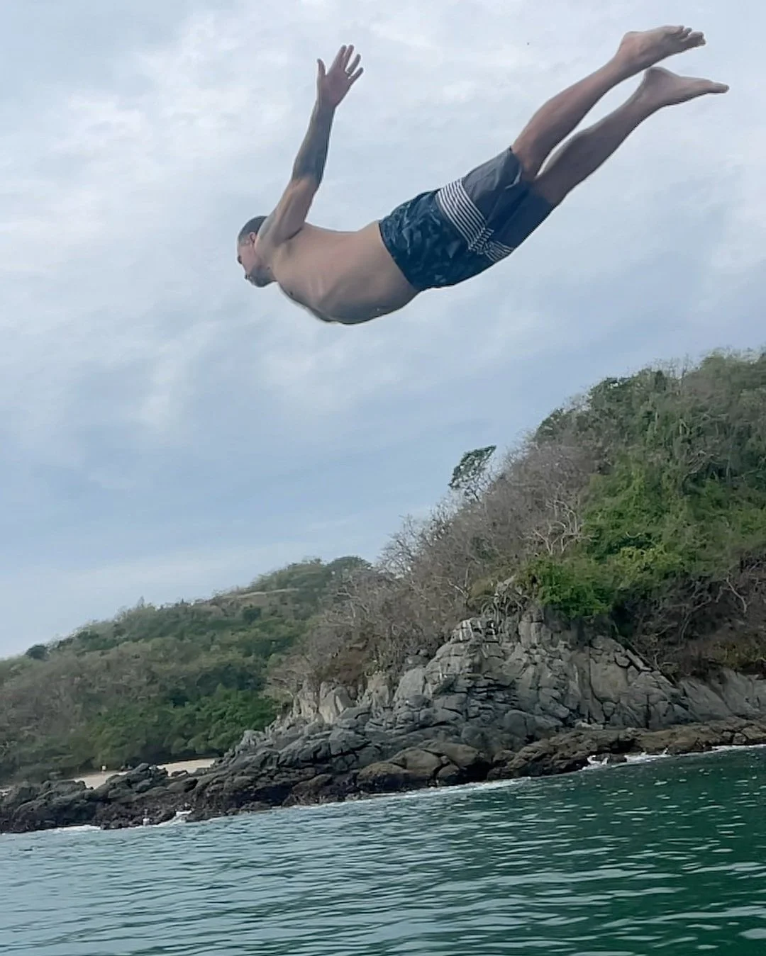 A man diving into the water from a rock ledge on a cloudy day at a beach with trees and rocks along the shoreline.