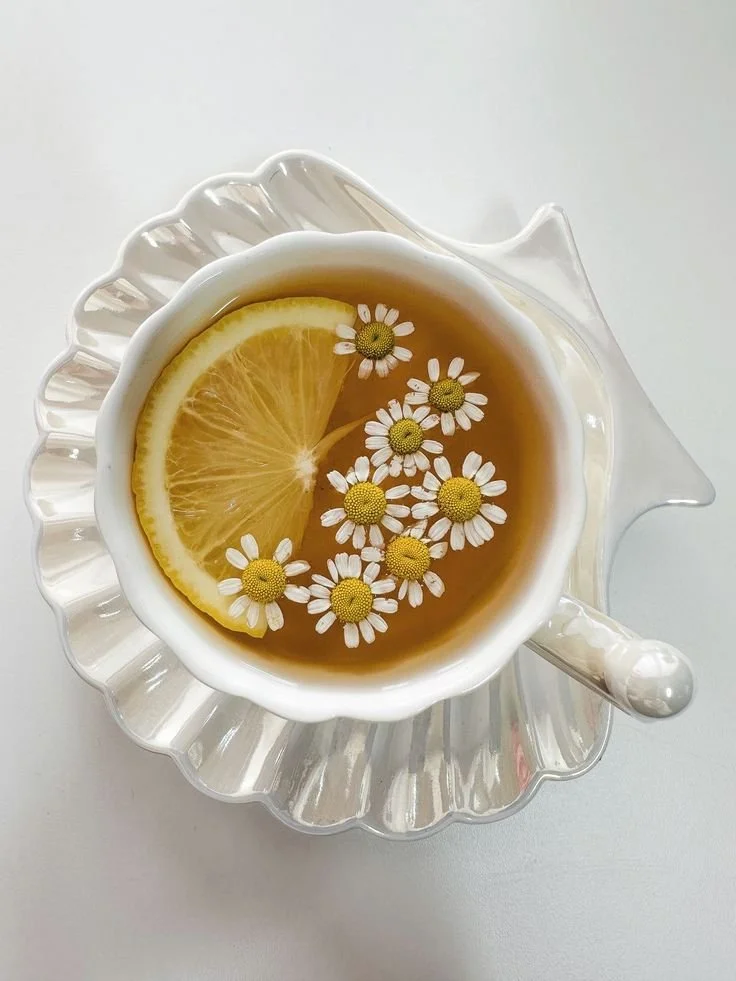 A white teacup with a scalloped edge and a small handle, filled with tea, garnished with a lemon slice and chamomile flowers, placed on a clear glass saucer with a ridged pattern.