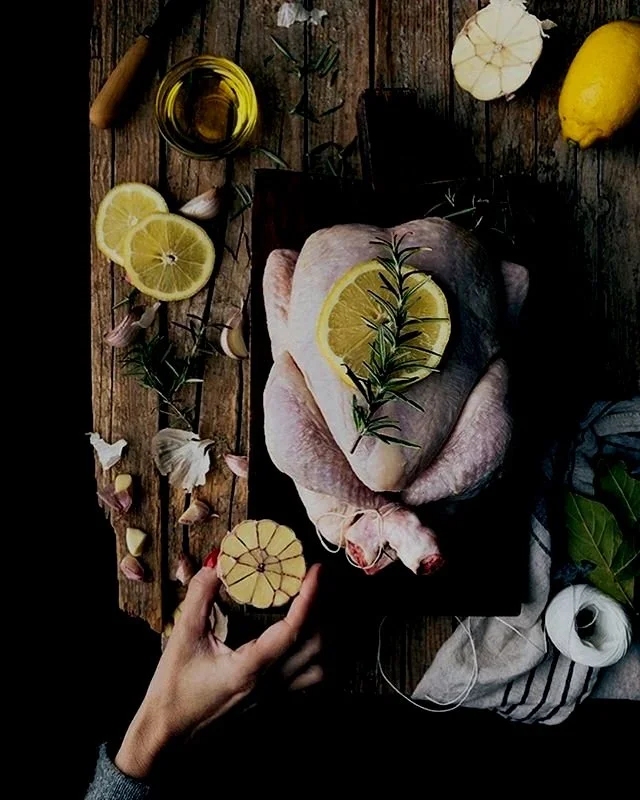 Raw whole chicken on a black cutting board topped with lemon slices and a sprig of rosemary, surrounded by lemon slices, garlic cloves, a lemon, a jar of oil, and kitchen towels on a rustic wooden table.