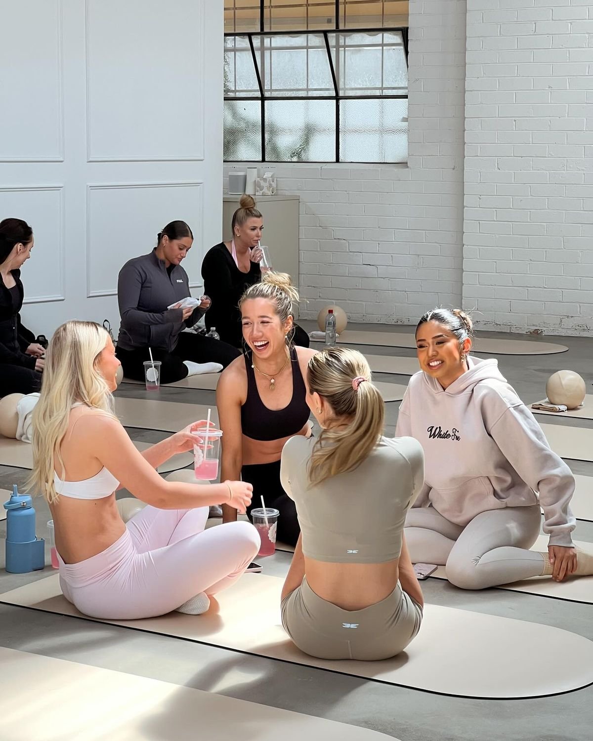 A group of women in workout clothes sitting on yoga mats, socializing and laughing during a fitness class in a bright, modern studio.