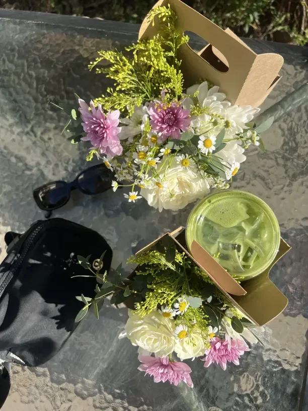 Two bouquets of white and pink flowers with green foliage on a glass table, each in a cardboard container. One contains a black camera with a strap next to it, and the other has a green iced drink in a plastic cup with a straw.
