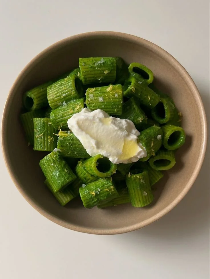 A beige bowl filled with cooked green pasta topped with a dollop of white ricotta cheese.
