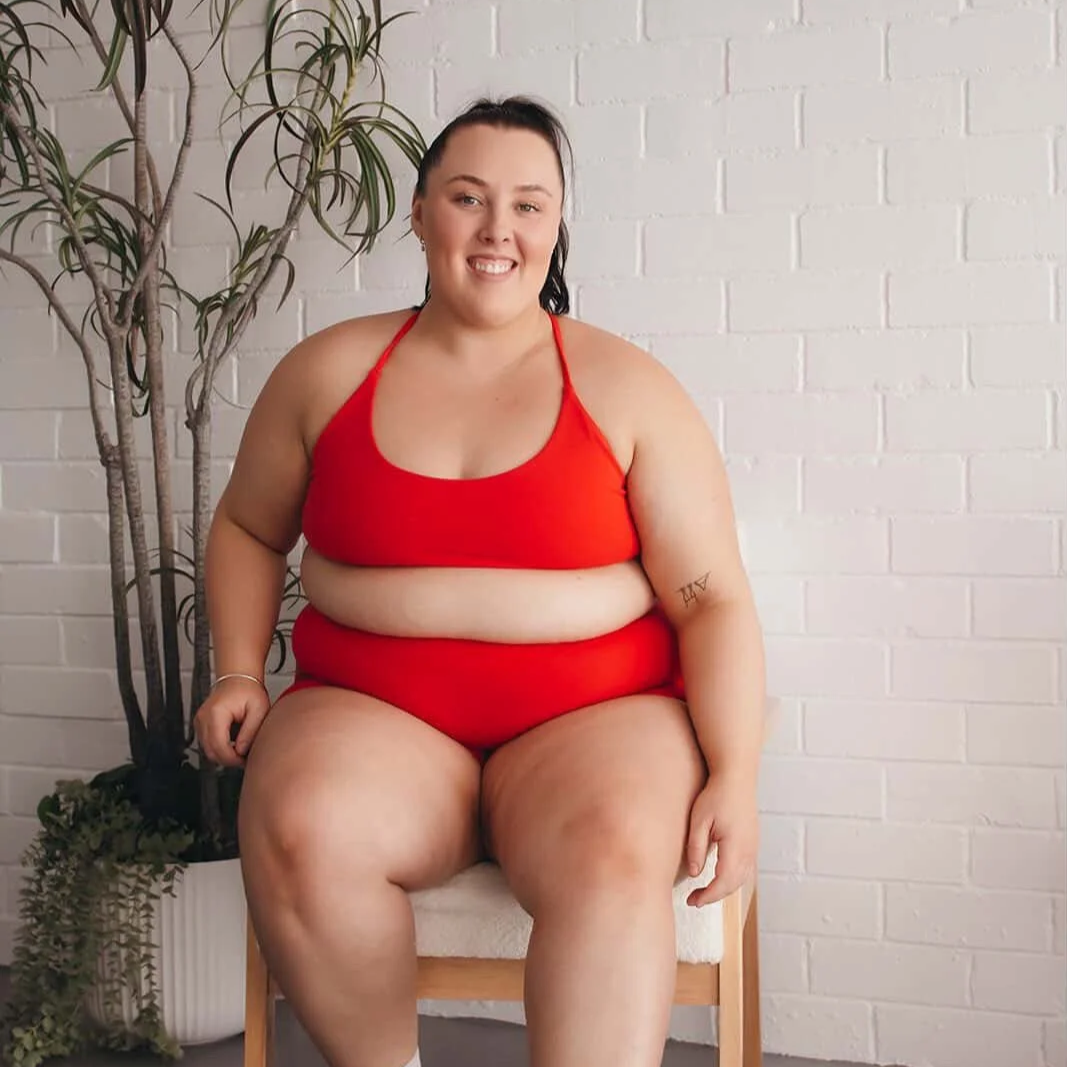 A smiling woman with dark hair in a red tank top and red shorts sitting on a chair against a white brick wall, with a tall plant to her left.