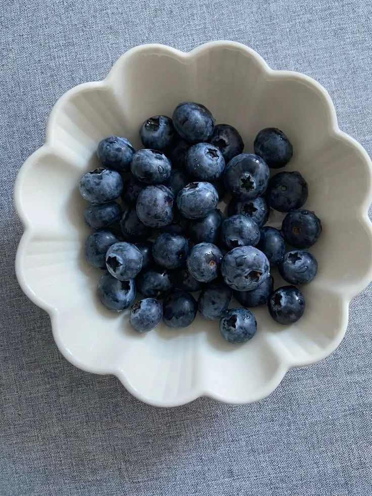 A white flower-shaped bowl filled with fresh blueberries on a gray fabric surface.