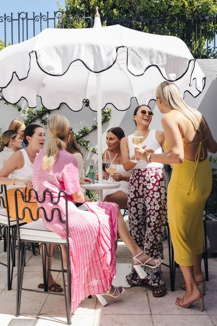 A group of women enjoying drinks at an outdoor patio with a white umbrella, bright sunlight, and leafy greenery in the background.