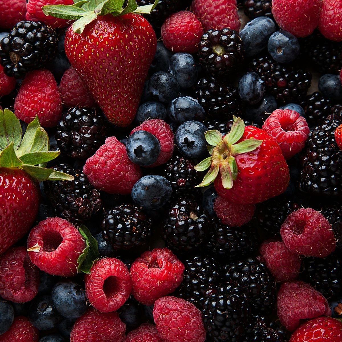 A close-up view of fresh strawberries, raspberries, blueberries, and blackberries, showing vibrant red and blue colors with green leaves on some strawberries.