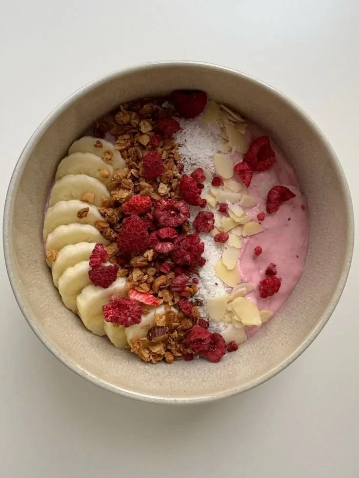 Bowl of yogurt topped with sliced banana, granola, raspberries, coconut flakes, and a berry-flavored sauce