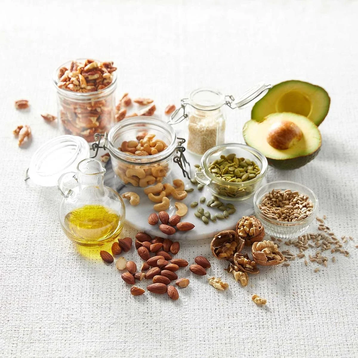A variety of nuts, seeds, and grains including almonds, cashews, walnuts, pumpkin seeds, sunflower seeds, and rice arranged in small glass jars and bowls, with halved avocado and a small bottle of olive oil on a white surface.