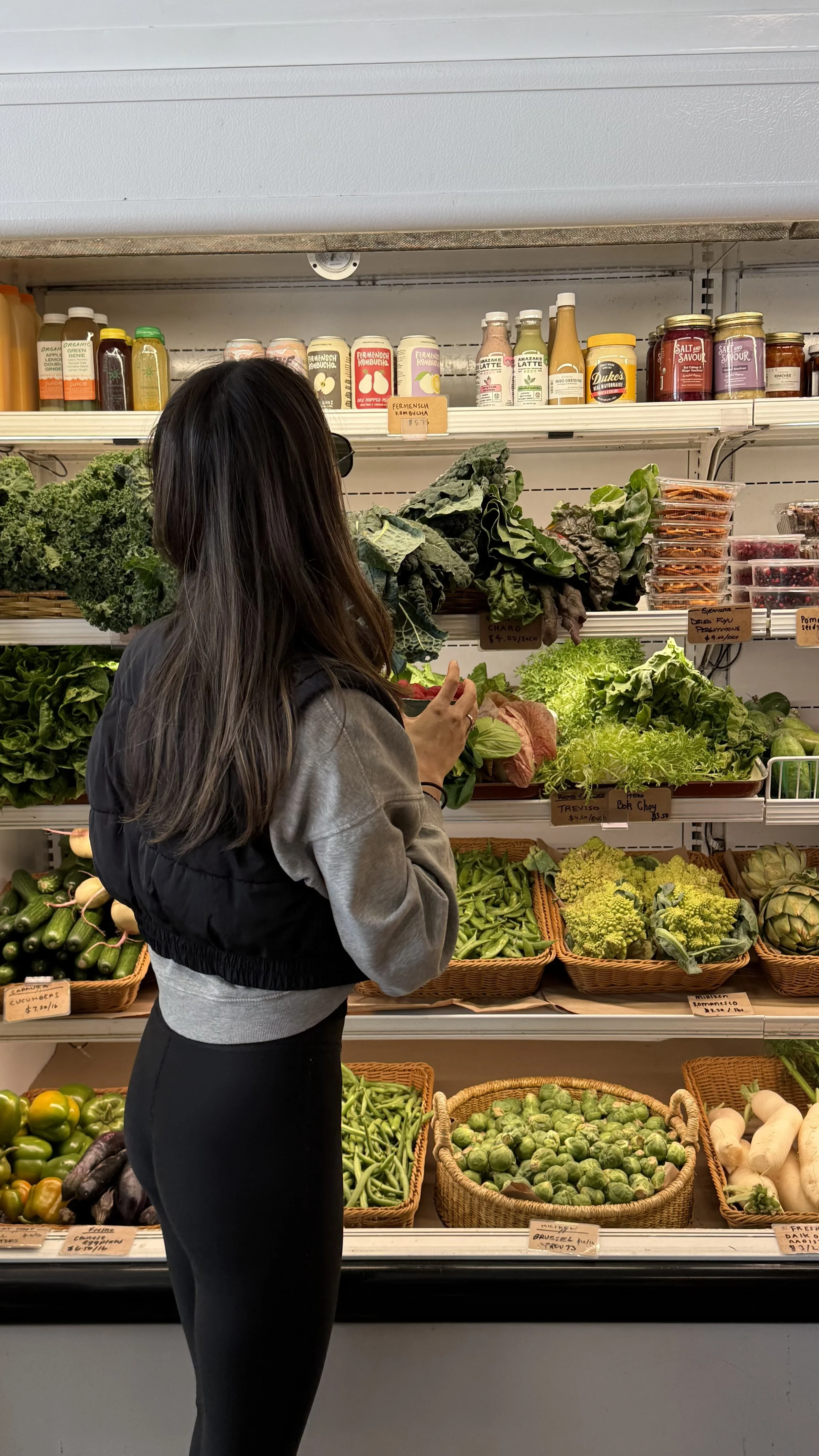 Woman shopping for fresh vegetables in a grocery store. She is facing the vegetable section with various greens, brussels sprouts, and other produce in baskets.