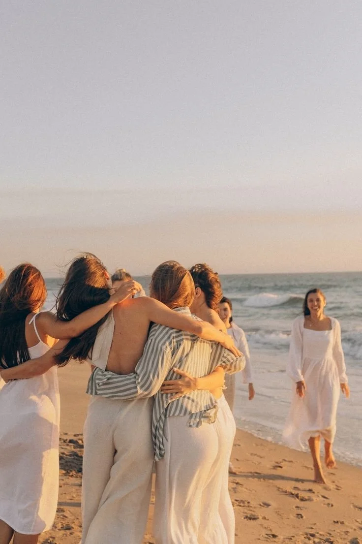 Group of women embracing on a beach at sunset, wearing white and striped clothing, with waves in the background.