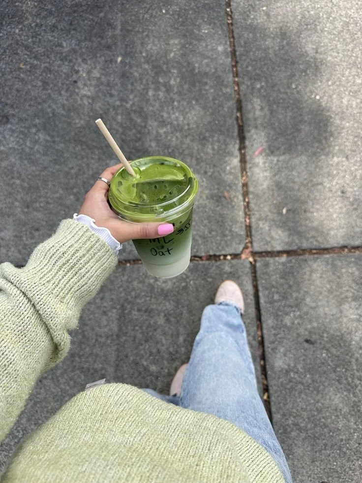 A person standing on a sidewalk holding a green and white bubble tea with a straw, wearing a light green sweater, blue jeans, and pink shoes.