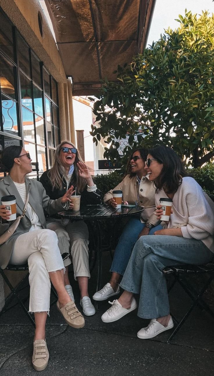 Five women sitting around a small black table outside a café, laughing and enjoying coffee, with a large green bush and building behind them.