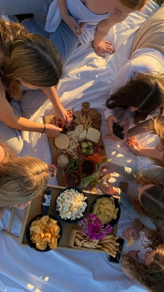 Group of people having a picnic with cheese, crackers, grapes, popcorn, chips, and vegetables on a table covered with white cloth.