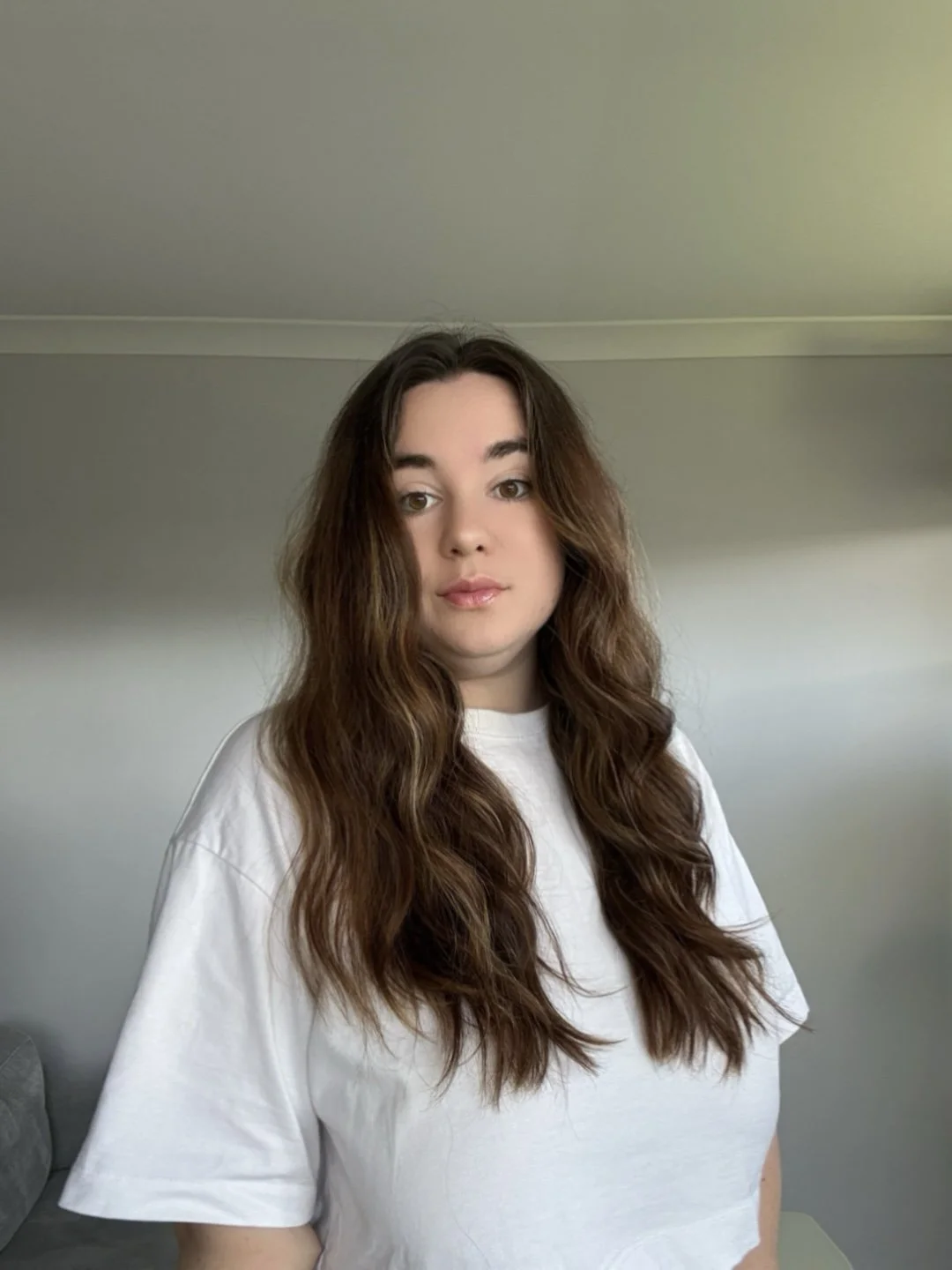 A woman with long wavy brown hair and light skin wearing a white t-shirt, standing in a room with plain walls.