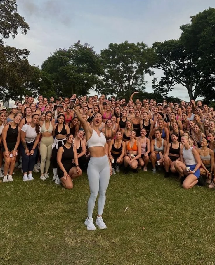 A large group of women in athletic wear gathered outdoors on grass, posing for a group photo, with one woman in front taking a selfie.