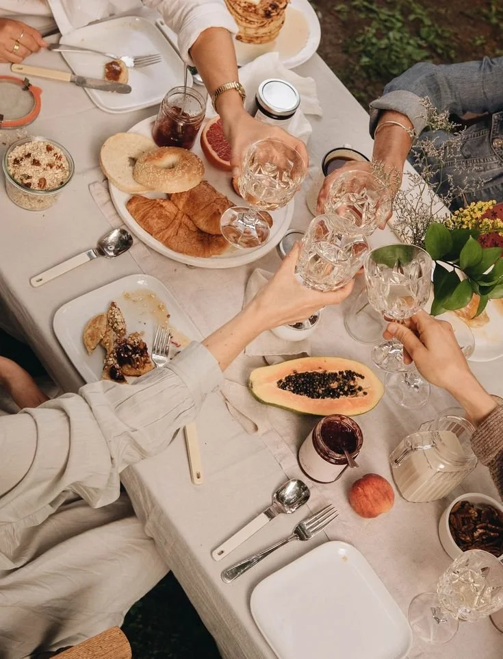 People clinking glasses of water at a table set with various breakfast foods, including a bagel, croissants, half a papaya with black seeds, and a bowl of cereal, during a meal outdoors.