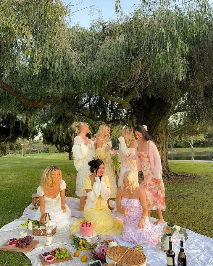 A group of women having a picnic outdoors under a large leafy tree, with some sitting on a white blanket on the grass and others standing. They are enjoying food, drinks, flowers, and each other's company.