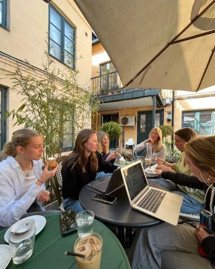 Six women sitting at a table outdoors, using laptops and mobile phones, with drinks and food on the table, in a cafe or restaurant setting.