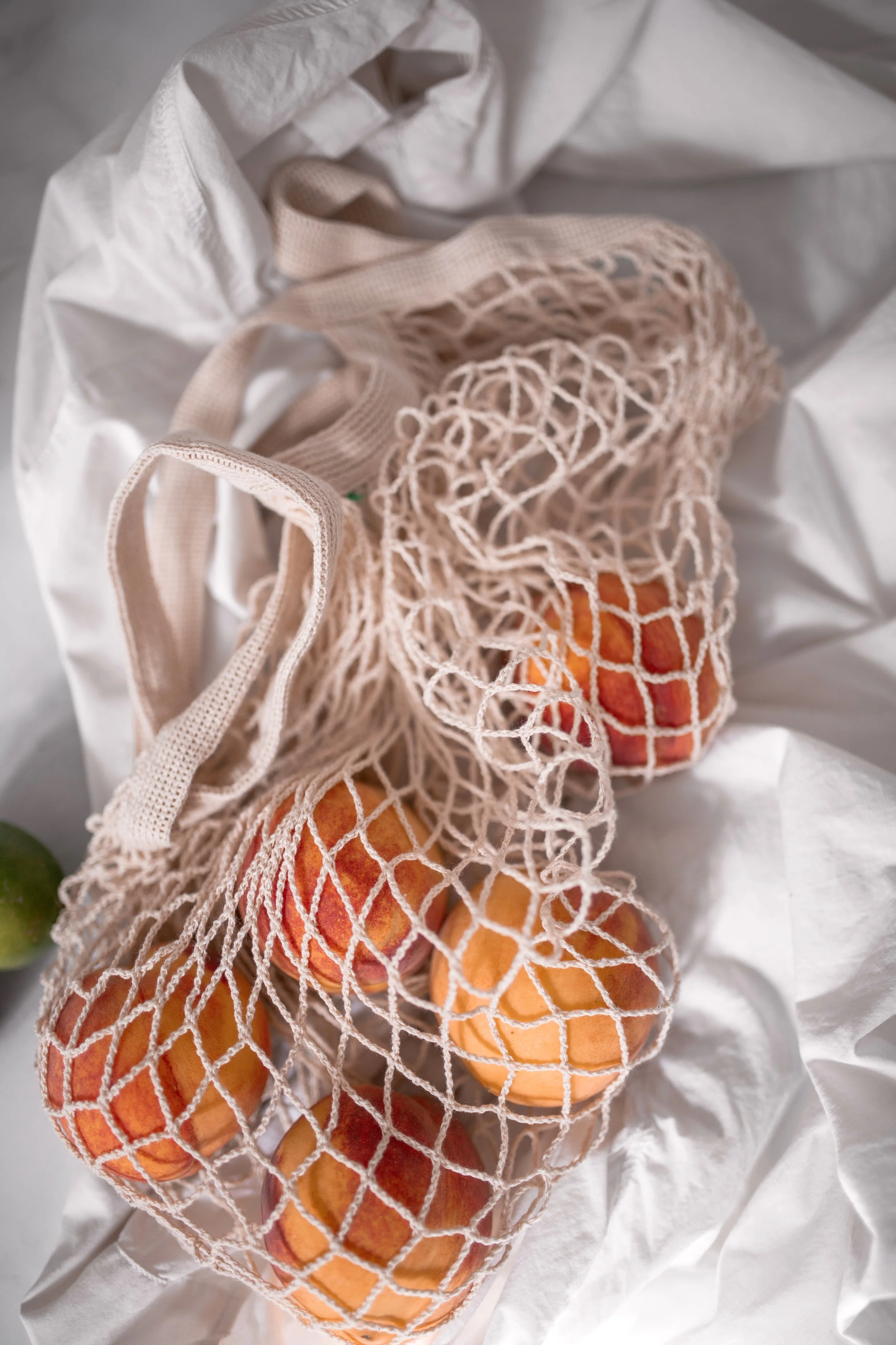 A beige reusable mesh shopping bag filled with peaches, resting on crumpled white cloth.