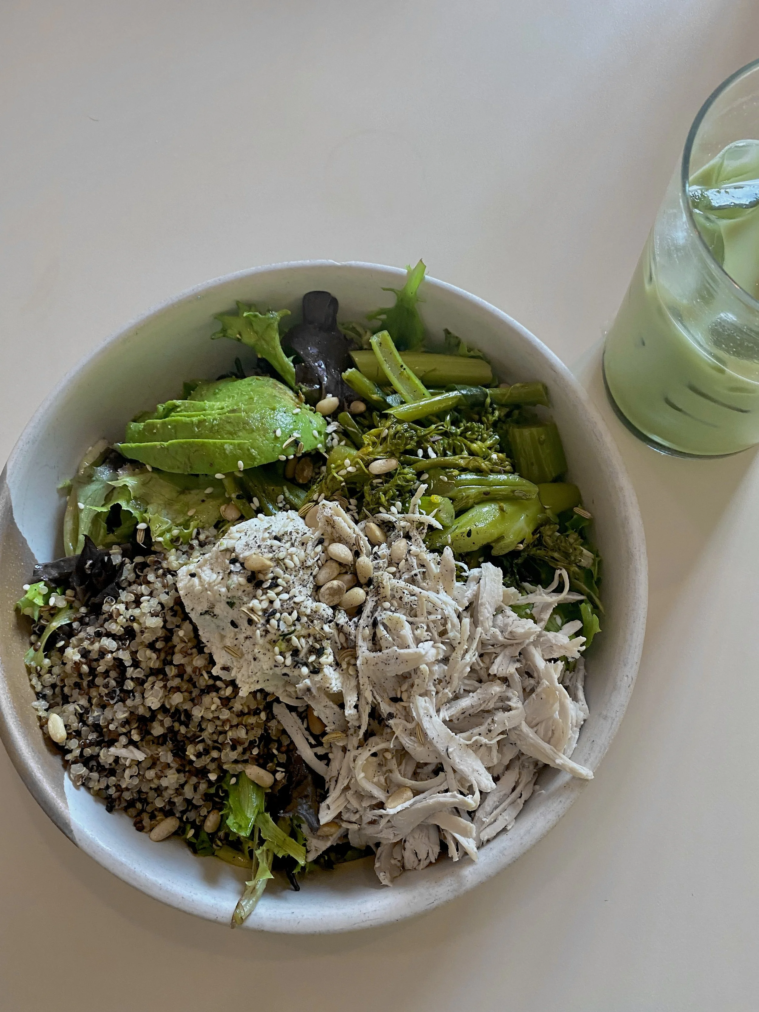 A bowl of salad with shredded chicken, avocado slices, mixed greens, seeds, and grains, accompanied by a glass of green smoothie.