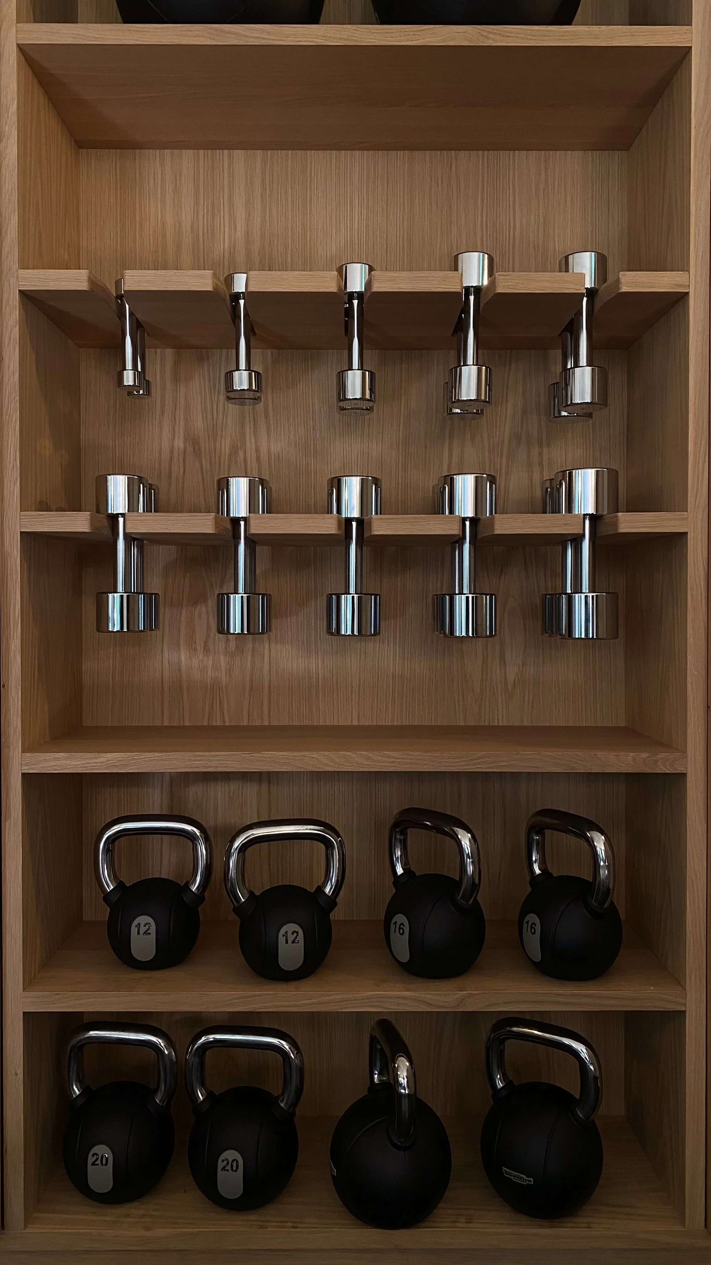 A wooden shelf containing six dumbbells on the top row and six kettlebells on the lower shelves, arranged by size.