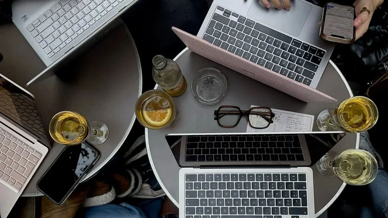 Two round tables with laptops, glasses of white wine, a smartphone, glasses, a beverage bottle, and a drink with lemon slices on both tables. One table also has a menu and a pink laptop case.