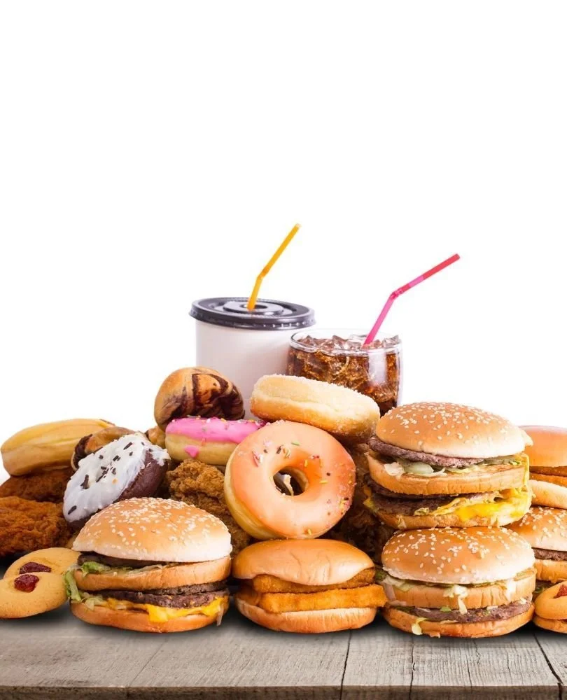 A variety of fast food items including cheeseburgers, donuts, cookies, chicken sandwiches, fries, and sodas displayed on a wooden surface.