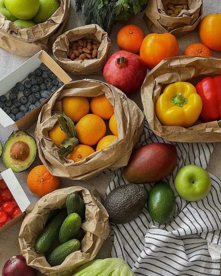 Assorted fresh fruits and vegetables including oranges, apples, bell peppers, avocados, cucumbers, blueberries, tomatoes, pomegranate, and leafy greens, arranged on a table with brown paper bags and a striped cloth.